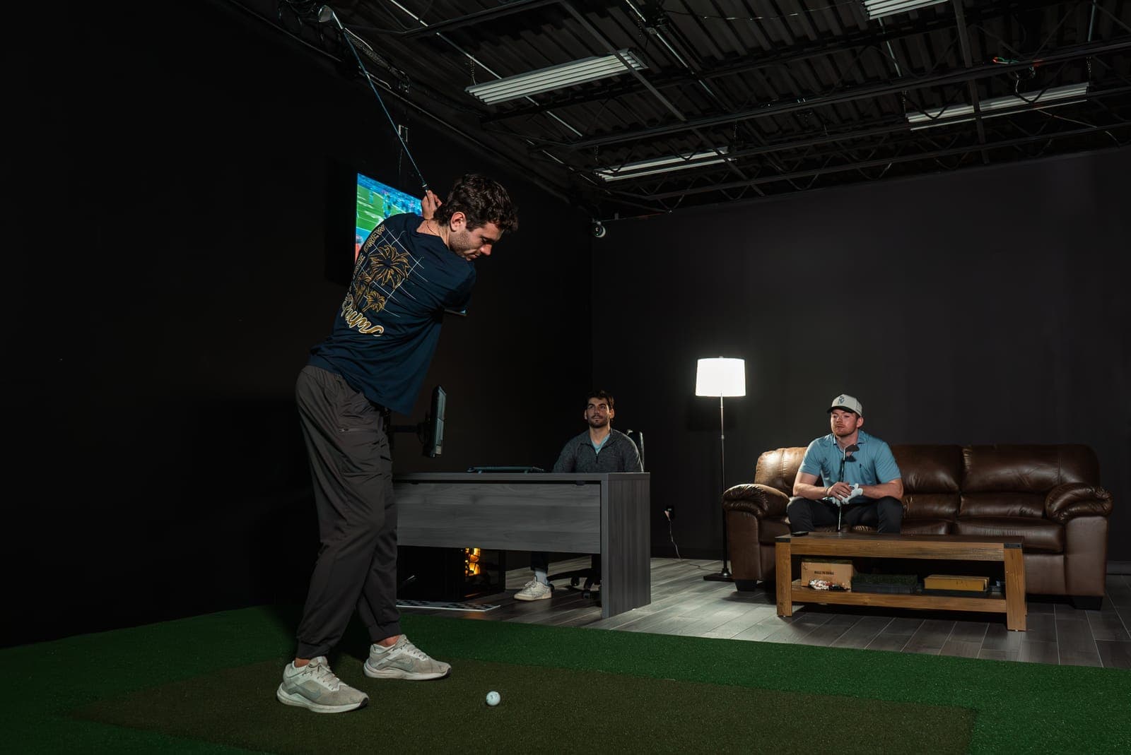 A golfer at the top of his backswing in a Franklin Square bay while two teammates watch from the couch and the desk.