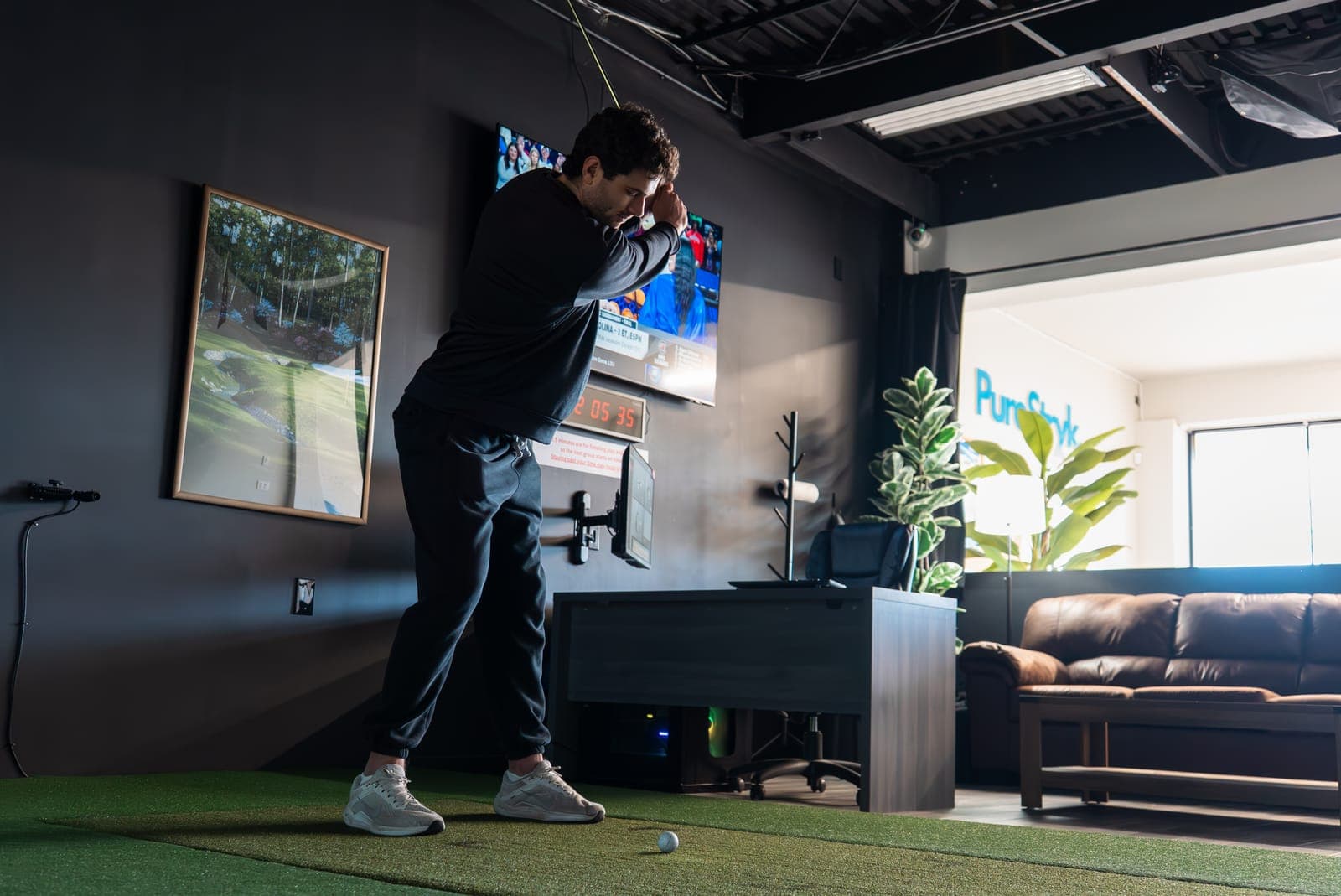 A golfer at the top of his backswing in a simulator bay, with a wall-mounted TV and a leather couch behind him.