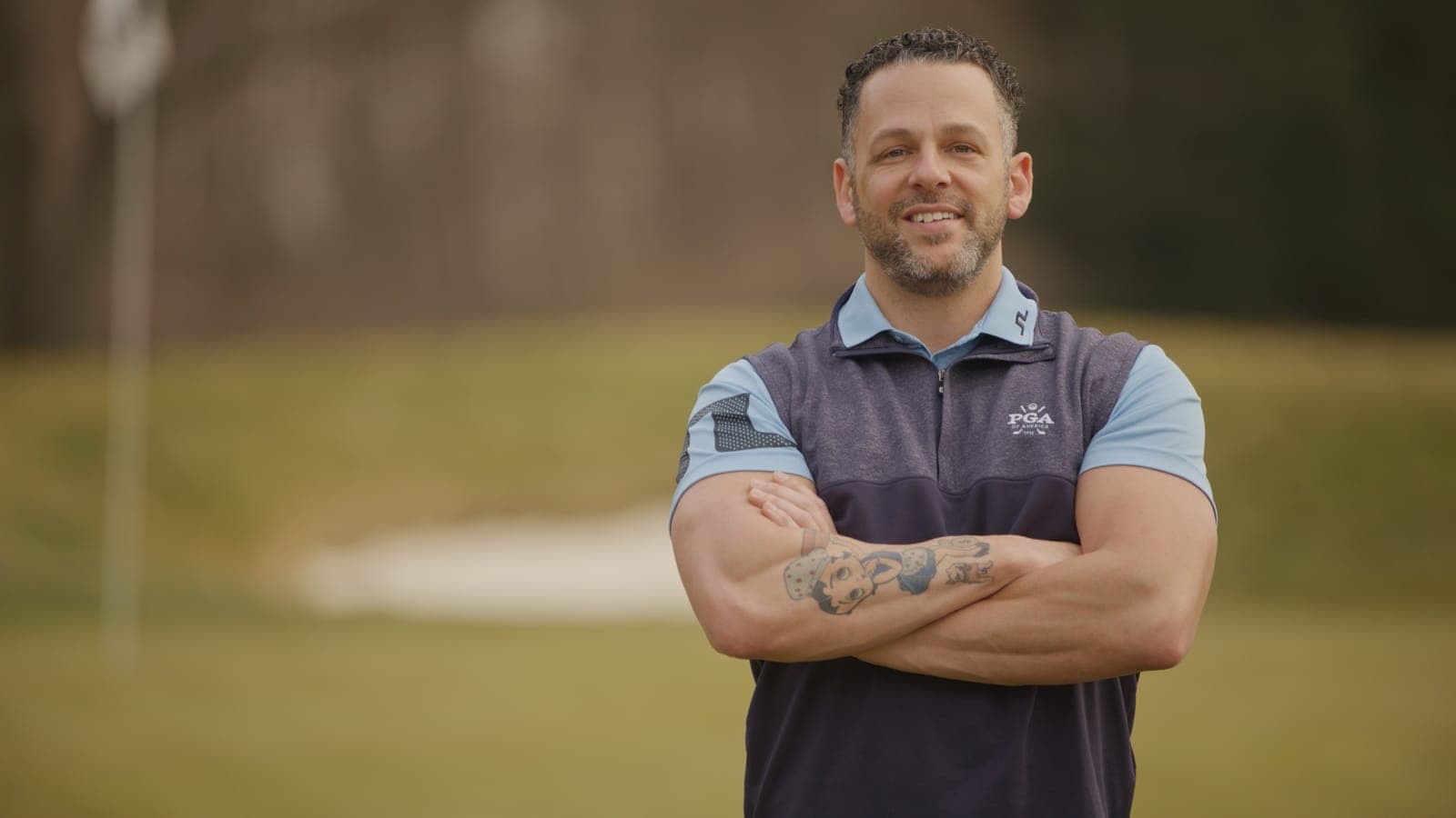 A teaching professional in a PGA of America polo, arms crossed, standing on a golf course.