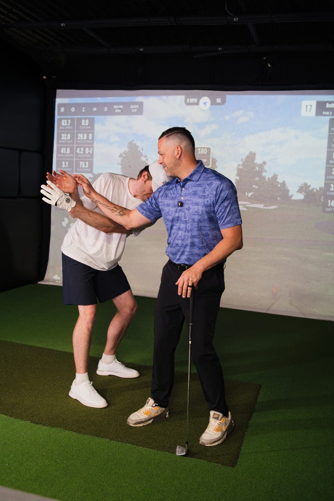An instructor adjusting a student's arms in front of a simulator screen showing shot data.
