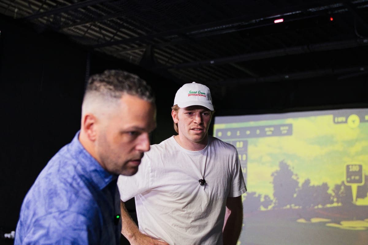 Two golfers in a simulator bay watching the screen, one wearing an Island Greens Invitational cap.