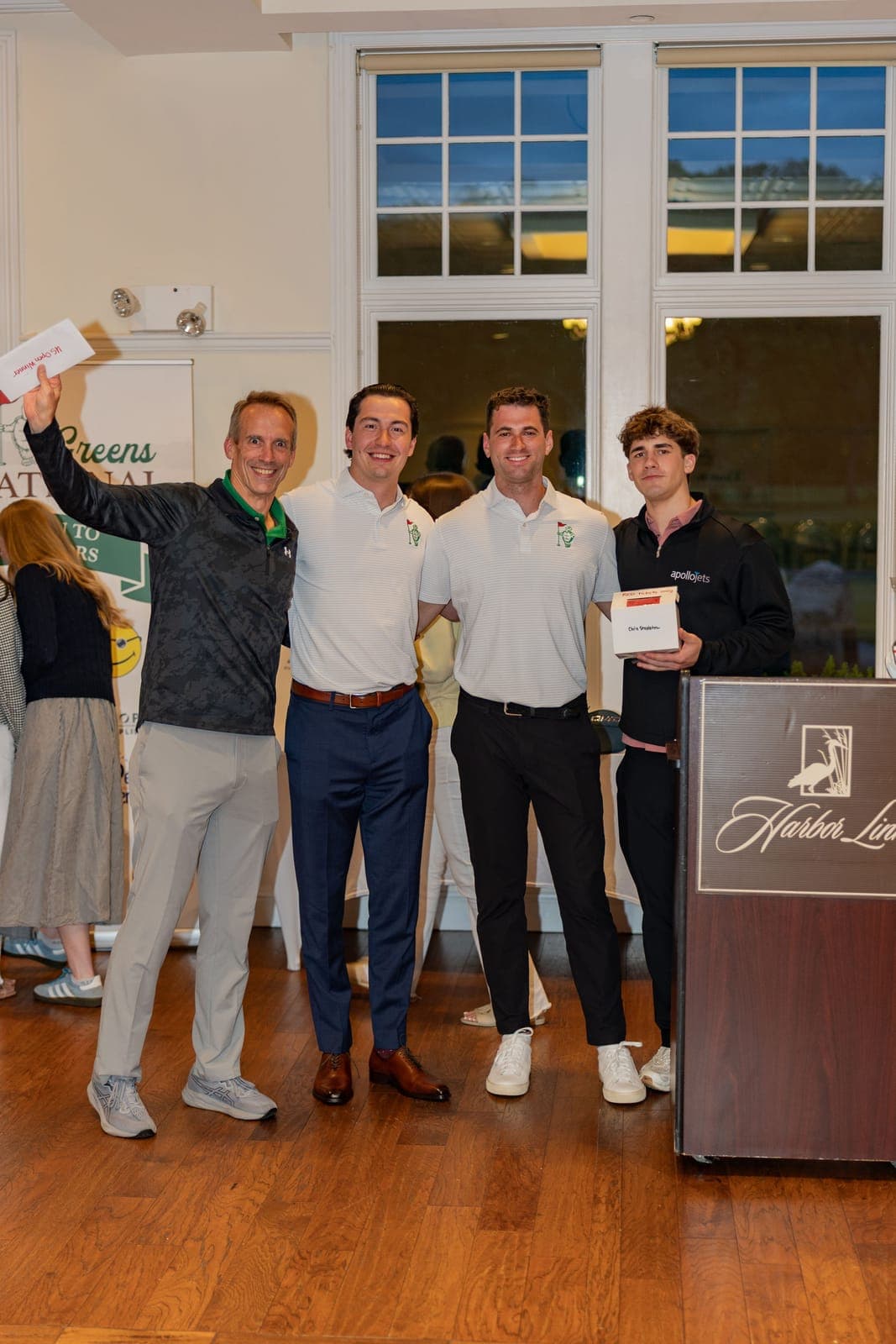 Four people at the lectern during the Island Greens Invitational awards, one holding up a raffle prize envelope.