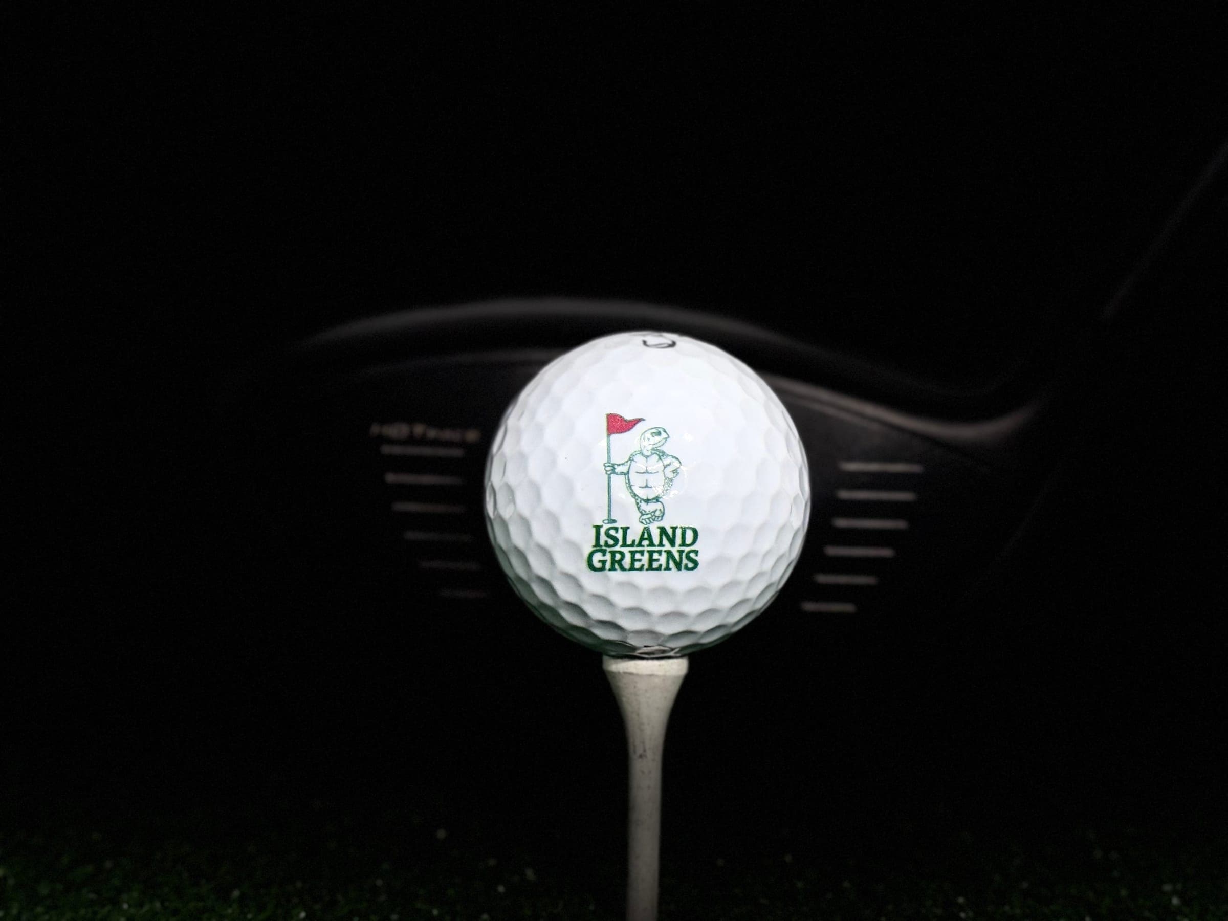 A white golf ball on a tee, printed with the Island Greens turtle and flag logo.